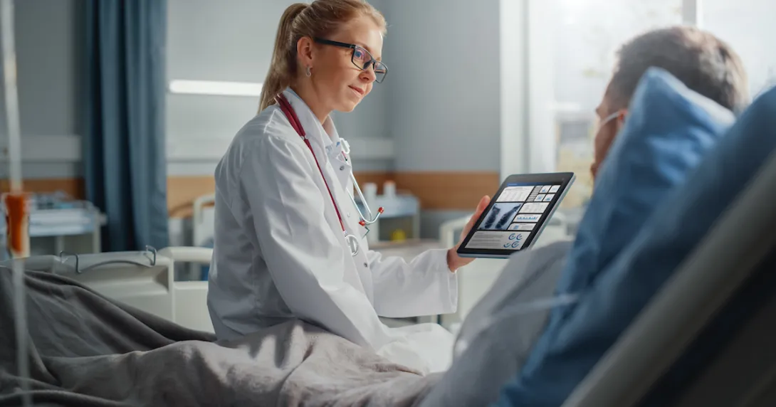 
  Healthcare provider sitting on a patient's hospital bed while holding a tablet showing diagnostic images of lungs
