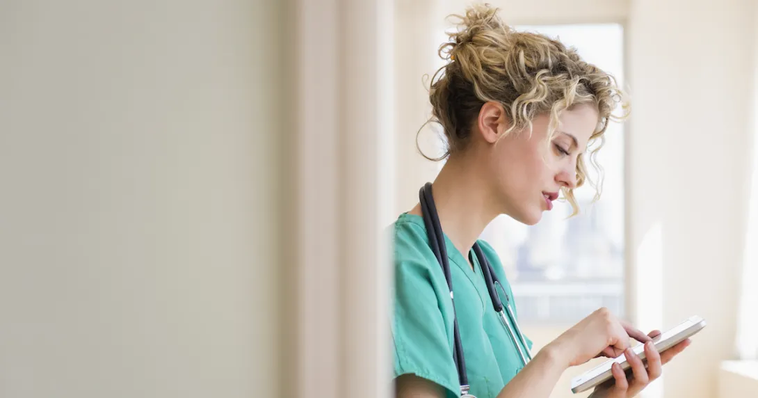 Healthcare provider in green scrubs wearing a stethoscope around their neck while looking at a tablet